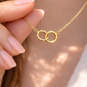 May include: A gold-colored necklace featuring two interlocking rings. One ring has a textured, twisted design, while the other is smooth. The necklace is suspended from a delicate chain, creating a simple yet elegant piece of jewelry.
