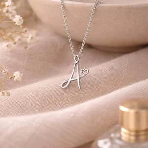 Könnte beinhalten: Eine silberfarbene Halskette mit einem Anhänger in Form des Buchstabens "A" und einem kleinen Herzdetail. Der Anhänger hängt an einer filigranen Kette. Der Hintergrund umfasst beigen Stoff, eine Keramikschale und eine Parfümflasche.