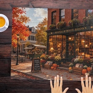 Puede incluir: Un rompecabezas que muestra una librer&iacute;a llamada Maple & Main Books con follaje oto&ntilde;al, calabazas y un carrito de caf&eacute;. Una pizarra dice Books Stories & Coffee Welcome. Una taza de t&eacute; blanca se encuentra sobre una superficie de madera oscura.