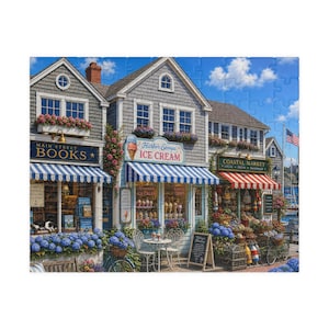 May include: A jigsaw puzzle depicting a coastal street scene with a bookstore, an ice cream shop, and a market. The buildings feature gray shingles, blue and white striped awnings, and flower boxes filled with pink and blue flowers under a blue sky.