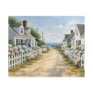 May include: A jigsaw puzzle featuring a watercolor painting of a coastal scene. White houses with black shutters line a sandy path leading to the ocean. Hydrangeas and a white picket fence frame the houses. An American flag hangs on one house.