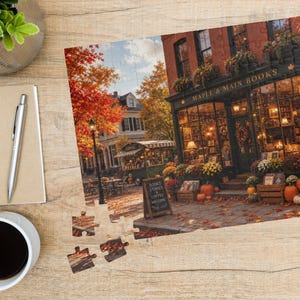 Puede incluir: Un rompecabezas que muestra una librer&iacute;a llamada Maple & Main Books con follaje oto&ntilde;al, calabazas y una escena callejera. El rompecabezas se exhibe sobre una superficie de madera junto a un cuaderno de espiral, un bol&iacute;grafo plateado y una taza blanca con caf&eacute; negro.