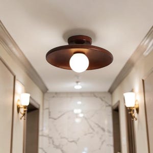 May include: A brown, circular ceiling light fixture with a white globe bulb. The light is centered in a hallway with white walls and marble-patterned accents. Two wall sconces with white shades are visible.