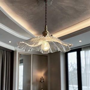 Puede incluir: Una lámpara colgante de cristal transparente con una pantalla plisada en forma de flor. La lámpara tiene una parte superior de color latón y un cable marrón. La bombilla es visible dentro de la pantalla. La luz cuelga de una lámpara de techo blanca.