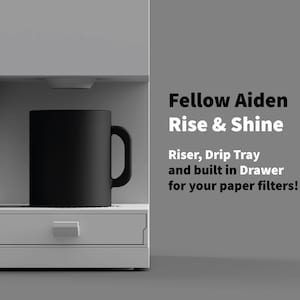 Könnte beinhalten: Ein moderner, minimalistischer Kaffeezubereiter mit einer schwarzen Tasse. Die Maschine ist weiß und hat eine eingebaute Schublade. Der Text lautet "Fellow Aiden Rise & Shine" und beschreibt Funktionen wie ein Podest und eine Abtropfschale.