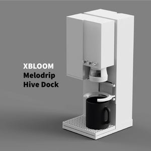 Könnte beinhalten: Eine weiße Kaffeemaschine mit einer schwarzen Tasse. Die Kaffeemaschine hat ein modernes Design mit dem Text "XBLOOM Melodrip Hive Dock" an der Seite. Die Tasse steht auf einem weißen Tablett.