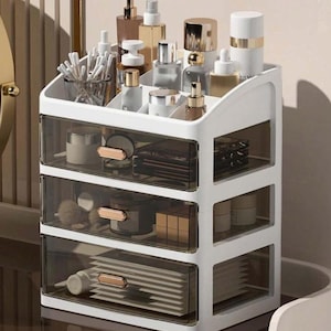 May include: A white plastic cosmetic organizer featuring three transparent dark-tinted drawers with gold-toned handles. The top section includes multiple open compartments holding various skincare bottles, jars, and a clear cup filled with cotton swabs.