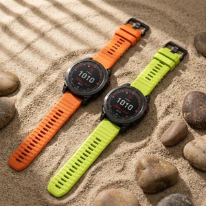 Puede incluir: Dos relojes inteligentes negros con el texto "1010" en la pantalla, cada uno con una correa de diferente color. Un reloj tiene una correa naranja y el otro una correa verde lima. Los relojes están sobre una superficie arenosa con pequeñas piedras.