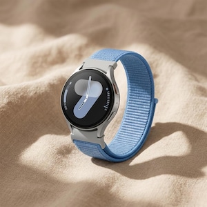 Op de afbeelding: Een zilveren smartwatch met een lichtblauwe stoffen band. De wijzerplaat toont een digitale klok met een lichtblauw en wit ontwerp. Het horloge ligt op een beige oppervlak.