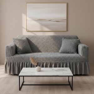 May include: A grey sofa with a textured cover and matching cushions. The sofa has a skirted base and is positioned beneath a neutral-toned painting. A white marble-topped coffee table with a black metal frame is in front of the sofa.
