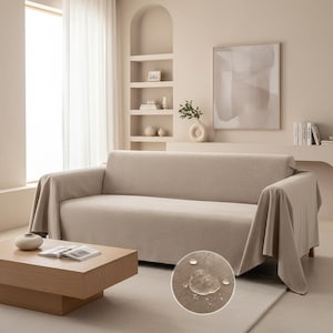 Puede incluir: Funda de sofá beige sobre un sofá de tres plazas. La funda tiene un aspecto texturizado y cuelga sobre los brazos. Una mesa de centro de madera con un libro y un jarrón decorativo están frente al sofá. Un primer plano muestra gotas de agua en la funda.