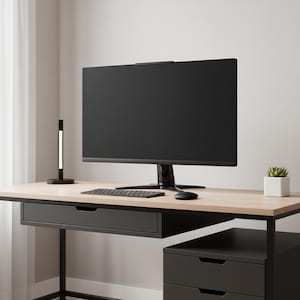 May include: A modern office desk with a large black computer monitor, keyboard, and mouse. A small desk lamp and a potted succulent are also on the desk. The desk has a light wood top and black metal frame with drawers.