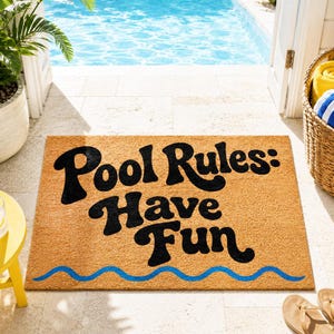 May include: A rectangular doormat with the words "Pool Rules: Have Fun" in black lettering. A blue wavy line is at the bottom. The mat is made of a natural fiber material and is placed near a pool.