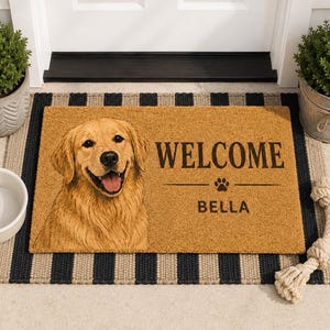 Op de afbeelding: Een rechthoekige beige kokosmat met een bedrukte afbeelding van een golden retriever aan de linkerkant. De tekst WELCOME staat in het zwart boven een pootafdruk-icoon, met de naam BELLA in zwarte letters daaronder gedrukt.