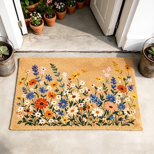 Può includere: Un zerbino rettangolare con sfondo beige e un colorato motivo floreale. I fiori includono margherite arancioni, blu, gialle e bianche e altri fiori selvatici. Il tappeto è posto davanti a una porta bianca.