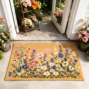 May include: A rectangular doormat with a floral design. The mat is tan with a border of colorful flowers in shades of pink, blue, white, and purple. The flowers are arranged in a row across the bottom of the mat.