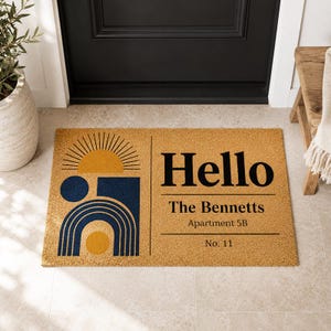 Könnte beinhalten: Eine rechteckige Fußmatte mit beigem Hintergrund zeigt ein geometrisches Design in Marineblau und Gelb, mit dem Wort "Hello" in großen schwarzen Buchstaben. Darunter steht "The Bennetts, Apartment 5B, No. 11."