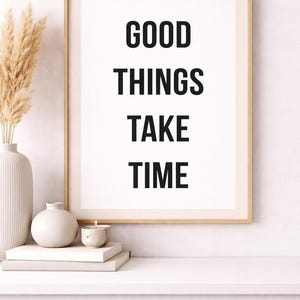 Könnte beinhalten: Gerahmter Druck mit dem Text „GOOD THINGS TAKE TIME“ in fetten schwarzen Buchstaben. Der Druck befindet sich in einem goldenen Rahmen und wird auf einem weißen Regal mit dekorativen Gegenständen ausgestellt, darunter eine Vase mit Pampasgras, eine kleine Kerze und Bücher.