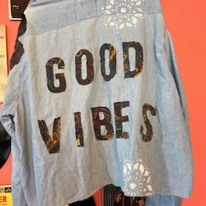 Könnte beinhalten: Hellblaues Jeanshemd mit den Worten "GOOD VIBES" auf dem Rücken in einem Paisley-Muster aufgenäht. Das Hemd hat weiße Blumenmuster und hängt an einer Wand mit Schallplatten.