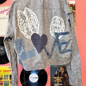 Könnte beinhalten: Ein graues Hemd mit Knöpfen, Aufnähern und dem Wort "LOVE" auf der Rückseite. Das "L" ist aus Jeansstoff, das "O" ist ein weißer Spitzenzirkel, das "V" ist ein Jeans-Patch und das "E" ist aus Jeansstoff. Ein dunkelblaues Herz befindet sich in der Mitte.