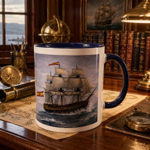 May include: White ceramic mug with a navy blue handle and rim. The mug features a detailed illustration of a tall ship with Spanish flags sailing on the ocean. The mug is set on a wooden desk with nautical decor.