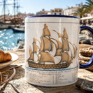 Pode incluir: Caneca de cerâmica branca com alça e borda azuis, com uma ilustração detalhada de um veleiro. A caneca tem texto em língua estrangeira na parte inferior. Um croissant, óculos de sol e corda náutica ao fundo.