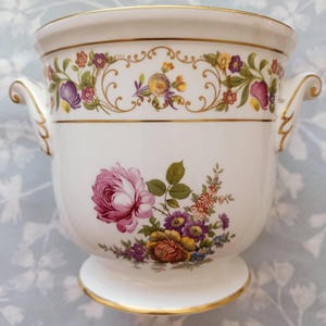 May include: A white porcelain cachepot featuring a gold-trimmed rim and base, two side handles, and a floral motif. The upper section displays a continuous garland of small colorful flowers, while the center features a larger bouquet of roses.