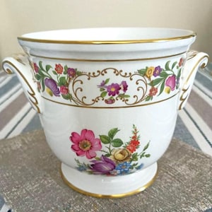 Könnte beinhalten: Weißer Keramik-Blumentopf mit goldenen Verzierungen und Griffen. Der Topf hat ein Blumendesign mit rosa, lila, gelben und blauen Blumen. Ein dekoratives goldenes Band mit floralen Akzenten umrundet den Topf.