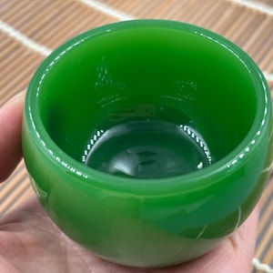 May include: A vibrant green glass bowl with a rounded shape. The bowl is held in a hand, showcasing its smooth, glossy surface. The interior reflects light, highlighting the depth of the color. The bowl is likely used for serving or display.