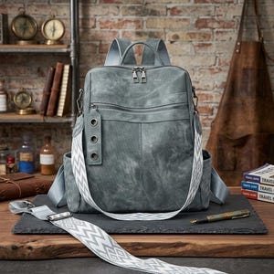 May include: A grey canvas backpack with a white and grey patterned shoulder strap. The backpack has a zippered main compartment and a front zippered pocket. The strap is adjustable. The bag is on a wooden surface.
