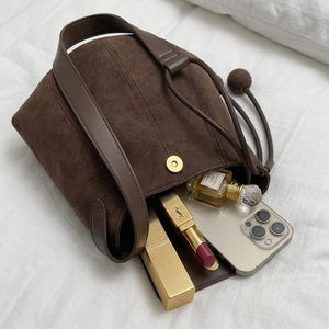 K&ouml;nnte beinhalten: Eine braune Wildlederhandtasche mit Schulterriemen und Kordelzugverschluss. Die Tasche ist offen und zeigt einen goldenen Lippenstift, ein Smartphone und eine Parf&uuml;mflasche. Die Tasche hat einen Magnetverschluss.