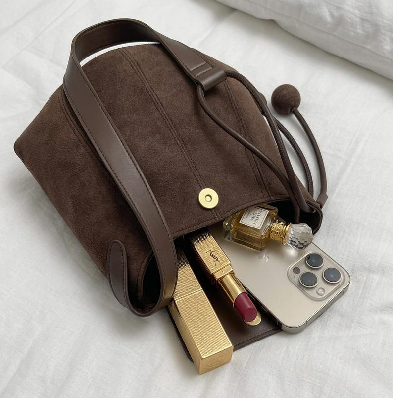 K&ouml;nnte beinhalten: Eine braune Wildlederhandtasche mit Schulterriemen und Kordelzugverschluss. Die Tasche ist offen und zeigt einen goldenen Lippenstift, ein Smartphone und eine Parf&uuml;mflasche. Die Tasche hat einen Magnetverschluss.