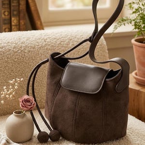 K&ouml;nnte beinhalten: Braune Wildleder-Umh&auml;ngetasche mit Klappenverschluss und langem Riemen. Die Tasche hat eine runde Form und eine dunkelbraune Lederklappe. Der Hintergrund umfasst eine Vase mit Blumen und einen kleinen Anh&auml;nger.