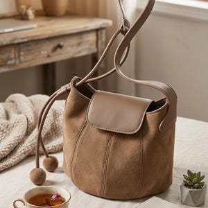 K&ouml;nnte beinhalten: Eine braune Wildlederhandtasche mit &Uuml;berschlag und Tragegriff. Die Tasche hat einen Kordelzug mit zwei dekorativen Kugelakzenten. Eine Tasse Tee, ein Notizbuch und ein Stift liegen ebenfalls auf dem Tisch.