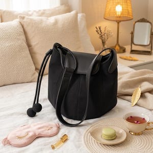 K&ouml;nnte beinhalten: Eine schwarze Bucket Bag mit Lederklappe und Schulterriemen liegt auf einem Bett. Eine rosa Augenmaske, eine Parf&uuml;mflasche, ein Macaron und eine Teetasse befinden sich ebenfalls auf dem Bett. Die Teetasse hat einen goldenen Rand und ist mit Tee gef&uuml;llt.