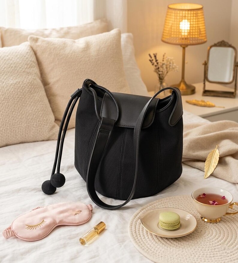 K&ouml;nnte beinhalten: Eine schwarze Bucket Bag mit Lederklappe und Schulterriemen liegt auf einem Bett. Eine rosa Augenmaske, eine Parf&uuml;mflasche, ein Macaron und eine Teetasse befinden sich ebenfalls auf dem Bett. Die Teetasse hat einen goldenen Rand und ist mit Tee gef&uuml;llt.