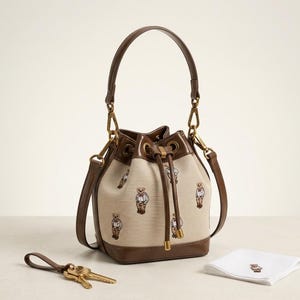 Può includere: Una borsa a secchiello beige con manico e finiture in pelle marrone, con ricami di orsetti. La borsa ha una chiusura con coulisse e dettagli dorati. Sono presenti anche un portachiavi abbinato e un fazzoletto bianco con un orsetto.