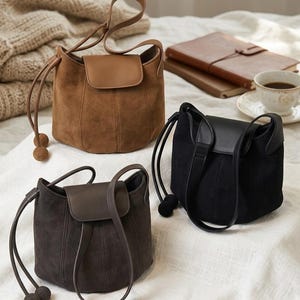 K&ouml;nnte beinhalten: Drei Wildleder-Eimertaschen in Braun, Dunkelbraun und Schwarz. Jede Tasche hat einen Klappenverschluss, Kordelzuggriffe mit dekorativen Kugelenden und vertikale Nahtdetails. Die Taschen werden auf einer wei&szlig;en Oberfl&auml;che pr&auml;sentiert.