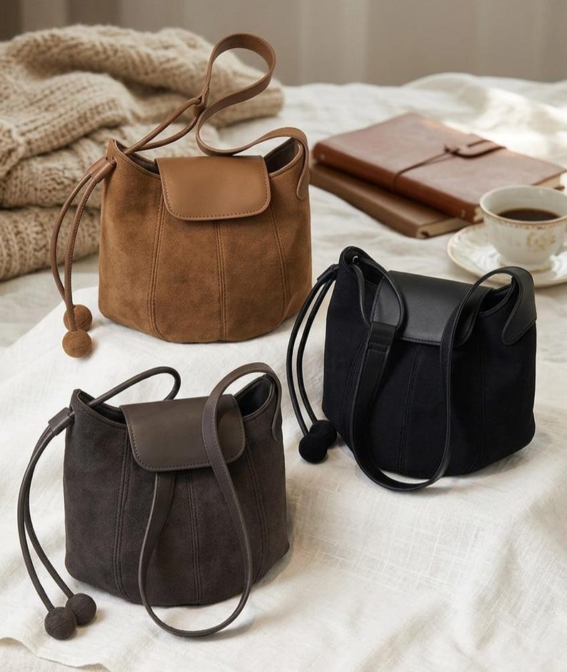 K&ouml;nnte beinhalten: Drei Wildleder-Eimertaschen in Braun, Dunkelbraun und Schwarz. Jede Tasche hat einen Klappenverschluss, Kordelzuggriffe mit dekorativen Kugelenden und vertikale Nahtdetails. Die Taschen werden auf einer wei&szlig;en Oberfl&auml;che pr&auml;sentiert.