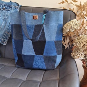 May include: A tote bag constructed from various shades of blue denim fabric patches. The bag features a rectangular label with the text DENIM DIVISION at the top centre and two matching denim fabric shoulder straps for carrying the item.