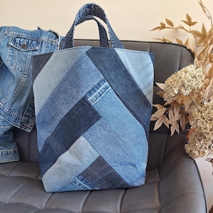 May include: A tote bag constructed from various shades of blue denim fabric arranged in a herringbone pattern. The bag features two matching denim handles at the top and a rectangular base, showcasing a patchwork design of light and dark blue denim.