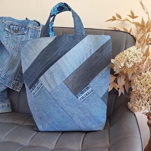May include: A tote bag constructed from various shades of blue denim fabric arranged in a diagonal patchwork pattern. The bag features two sturdy denim handles and an interior lining visible at the top with a blue and white patterned design.