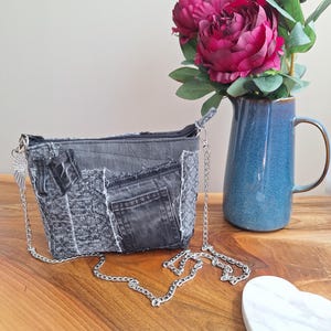 May include: A small crossbody bag crafted from repurposed grey denim fabric with a patchwork design, featuring frayed edges, a visible pocket, a top zipper closure, and a silver-toned metal chain strap adorned with a small wing-shaped charm.