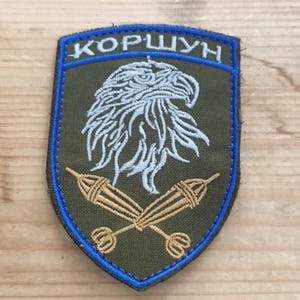 以下が含まれることがあります： オリーブグリーンの盾型刺繍ワッペン。白い鷲の頭のグラフィックと、その下に交差したベージュの矢が描かれています。ワッペンには鮮やかなブルーの縁取りがあり、上部にはキリル文字で「KOPШYH」と白く刺繍されています。