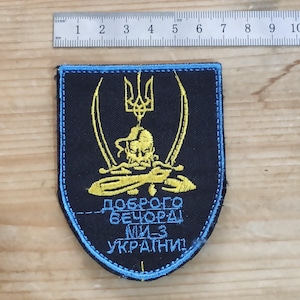 以下が含まれることがあります： 盾形の黒い刺繍ワッペンで、縁は水色です。黄色い三叉のエンブレムの下に、2本の剣を持った様式化された人物と、水色のキリル文字「ДОБРОГО ВЕЧОРА! МИ З УКРАЇНИ!」が描かれています。金属製の定規で幅が9cmであることが示されています。