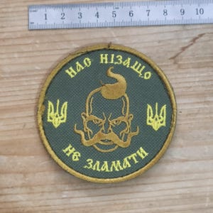 May include: A circular olive green embroidered patch featuring a yellow Cossack figure with a traditional hairstyle and moustache, flanked by two yellow trident symbols. The text "НАС НІЗАЩО НЕ ЗЛАМАТИ" is stitched in yellow. A metal ruler shows a 10 cm width.