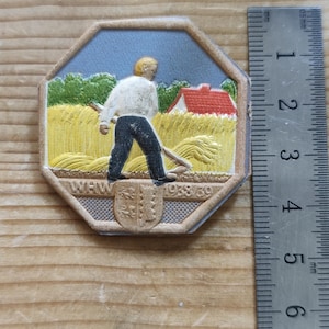 Peut inclure: Un badge octogonal présentant un relief d'une personne récoltant un champ de blé jaune avec une faux, une maison au toit rouge et des arbres verts. Le texte WHW 1938/39 est visible au-dessus d'un blason. Une règle montre 4 cm de largeur.