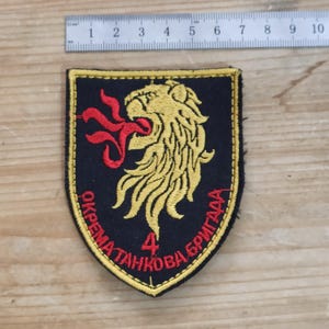 Peut inclure: Un écusson brodé en forme de bouclier représentant une tête de lion jaune crachant des flammes rouges sur fond noir. L'écusson porte le texte rouge "OKREMATANKOVABRYGADA" et le chiffre "4" en rouge. Une règle métallique indique 10 cm de large.