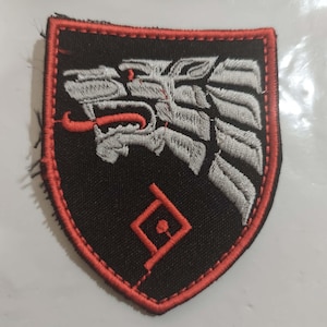 Peut inclure: Un écusson brodé en forme de bouclier avec un fond noir et une bordure rouge. Le motif représente une tête de loup argentée stylisée de profil avec une langue rouge, placée au-dessus d'un symbole géométrique rouge composé d'un losange et d'une ligne.