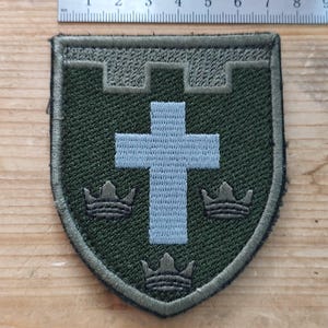 Op de afbeelding: Een olijfgroene geborduurde schildvormige patch met een lichtblauw kruis in het midden en drie donkere kronen eronder. Een metalen liniaal boven de patch laat zien dat deze een breedte heeft van ongeveer 6,5 cm.